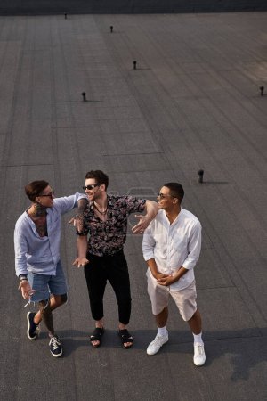 Téléchargez les photos : Trois amis multiraciaux joyeux en vêtements vibrants posant sur le toit et se regardant - en image libre de droit