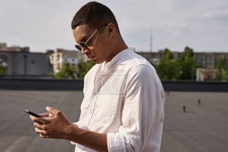 gut aussehender junger afrikanisch-amerikanischer Mann in Freizeitkleidung mit Sonnenbrille beim Blick auf das Smartphone