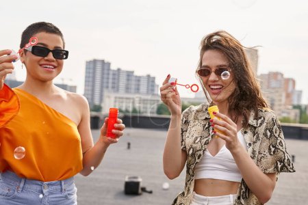 attraktive, fröhliche Frauen in lebendigen, lässigen Outfits mit Sonnenbrille, die Seifenblasen auf dem Dach bläst