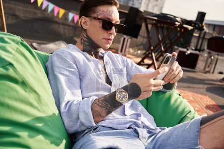 joven guapo con tatuajes y gafas de sol relajante durante la fiesta y mirando el teléfono inteligente