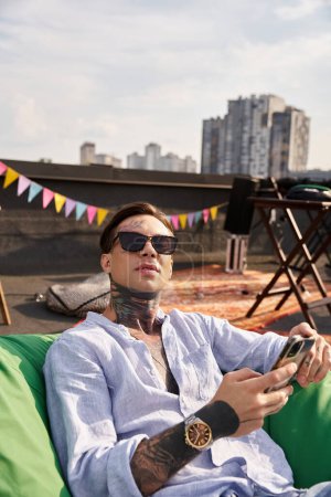fröhlicher gutaussehender Mann mit Tattoos und Sonnenbrille entspannt mit Handy in der Hand und blickt in die Kamera