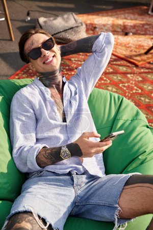 joyous attractive man with tattoos and sunglasses relaxing with phone in hand and looking at camera