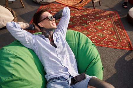 appealing cheerful man in vivid attire with sunglasses and tattoos relaxing on roof and looking away