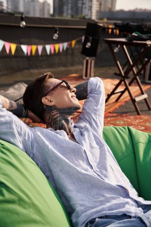 attraktiver fröhlicher Mann in lässiger Kleidung mit Sonnenbrille und Tattoos entspannt auf dem Dach und schaut weg