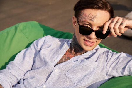 joyous handsome man in casual clothes with tattoos and stylish sunglasses smiling at camera on roof