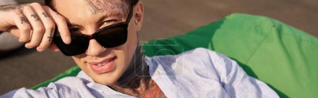 fröhlicher junger Mann mit Tattoos und stylischer Sonnenbrille, der auf dem Dach in die Kamera lächelt, Banner