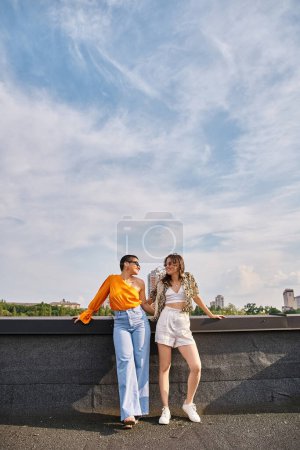 two cheerful beautiful women in vibrant clothes with sunglasses posing actively on rooftop