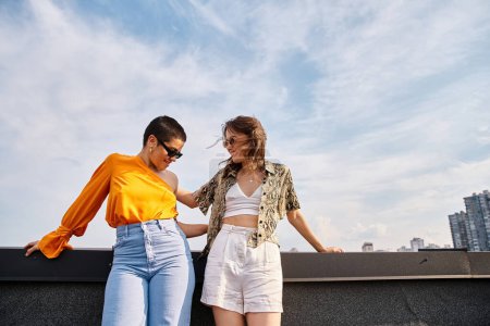 two joyful attractive women in stylish casual attires with sunglasses posing on rooftop together