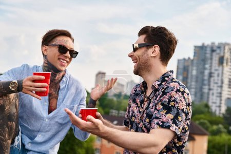 two cheerful men in casual attire with stylish sunglasses talking and holding red cups with drinks