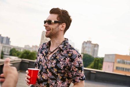 fröhlicher gutaussehender Mann mit stylischer Sonnenbrille, rote Tasse mit Getränk in der Hand und auf dem Dach wegschauend