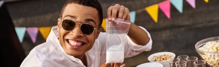 alegre hombre afroamericano con gafas de sol sosteniendo bolsa de sal y sonriendo a la cámara, pancarta