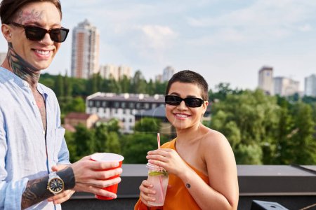 Téléchargez les photos : Joyeux homme et femme en tenue vive avec des lunettes de soleil boire à la fête sur le toit et sourire à la caméra - en image libre de droit