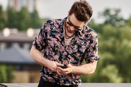 fröhlicher gutaussehender Mann mit Sonnenbrille in lebendiger Kleidung, der auf dem Dach auf sein Handy schaut