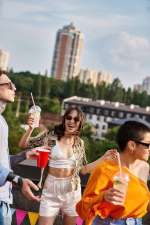 fröhliche multirassische junge Leute in lebendiger Kleidung, die auf dem Dach feiern und Cocktails trinken