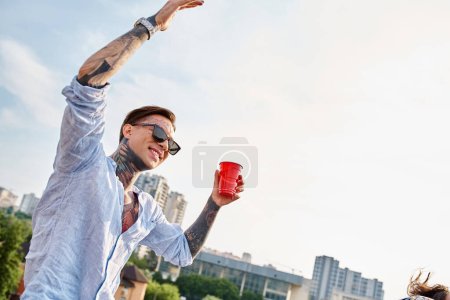 good looking cheerful man with tattoos and stylish sunglasses holding red cup at rooftop party