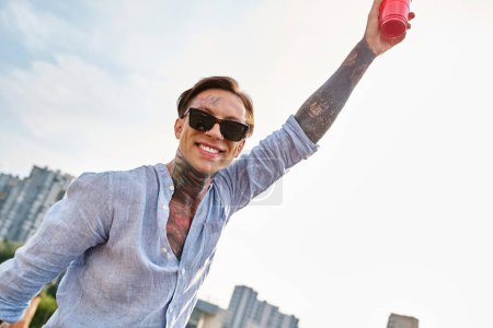 schöner fröhlicher Mann mit Tattoos und trendiger Sonnenbrille, der mit roter Tasse posiert und in die Kamera lächelt