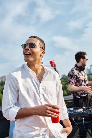 fröhlicher afrikanisch-amerikanischer Mann mit stylischer Sonnenbrille und roter Tasse mit Drink bei Dachparty