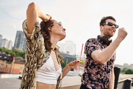 joyous woman with long hair and stylish sunglasses holding cocktail next to DJ at rooftop party