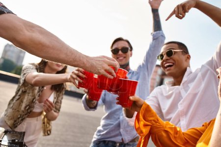 Téléchargez les photos : Jeunes amis multiraciaux joyeux accrochant leurs tasses rouges à côté de l'équipement DJ à la fête sur le toit - en image libre de droit