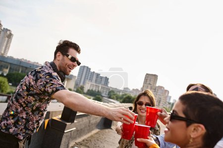 Téléchargez les photos : Jeunes amis joyeux divers claquant leurs tasses rouges à côté de l'équipement DJ à la fête sur le toit - en image libre de droit