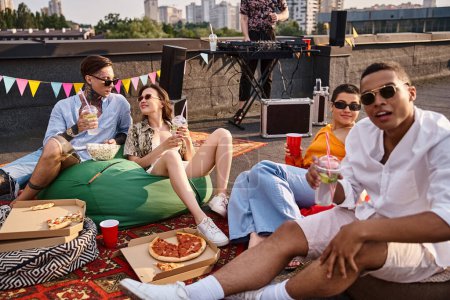 joyful multicultural friends with glasses sitting on rooftop with cocktails and pizza net to DJ