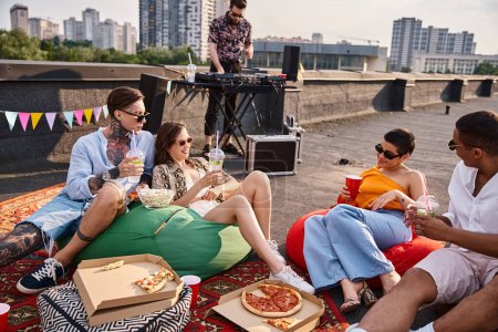 cheerful interracial friends with glasses sitting on rooftop with cocktails and pizza net to DJ