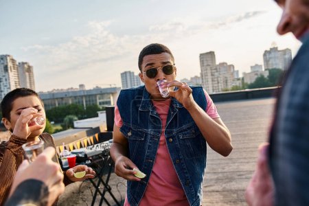 Téléchargez les photos : Homme afro-américain joyeux en gilet boire de la tequila avec de la chaux et du sel à côté de ses amis à la fête - en image libre de droit