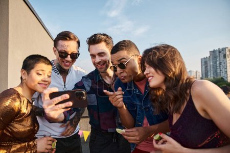 multiracial joyful people in casual clothes watching photos at smartphone at rooftop party
