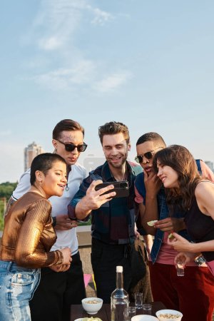 Multikulturelle junge Leute in lässiger Kleidung, die auf einer Dachparty Fotos auf dem Smartphone ansehen