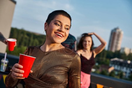 joyeuse femme aux cheveux courts avec bretelles tenant tasse rouge avec boisson avec son ami flou sur fond