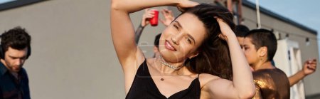 focus on cheerful woman looking at camera at rooftop party with her diverse blurred friends, banner