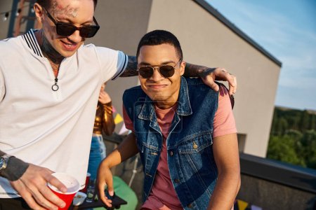 fröhliche multikulturelle Männer in lebhaften Freizeitanzügen, Cocktails in der Hand und auf der Party in die Kamera starrend