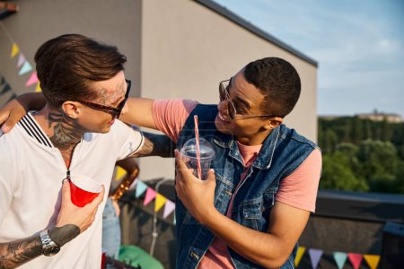 attractive joyous multicultural men holding cocktails and looking at each other at rooftop party