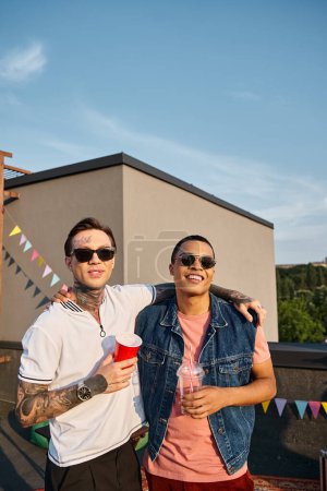 alegres hombres multiculturales en vibrantes trajes urbanos celebrando cócteles y mirando a la cámara en la fiesta