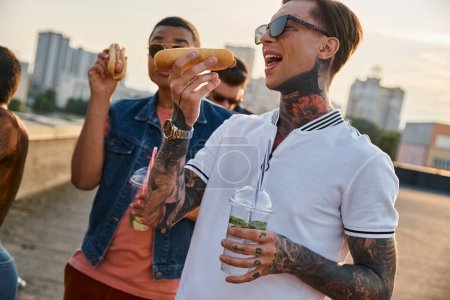 freudige multirassische Freunde in urbanen lebendigen Outfits genießen köstliche Hot Dogs auf dem Dach einer Party