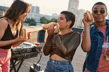 gut aussehende fröhliche interrassische Freunde in lässigen Outfits essen leckere Hot Dogs auf einer Dachparty