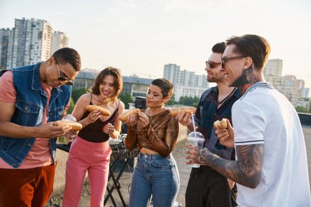 good looking jolly multicultural friends in urban outfits eating tasty hot dogs at rooftop party