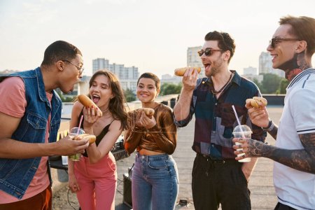 gut aussehende fröhliche diverse Freunde in lässigen Outfits essen leckere Hot Dogs auf der Dachparty