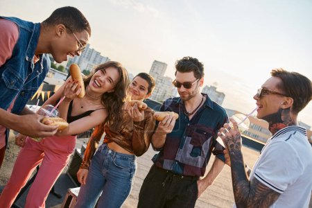 cheerful multicultural people in vivid outfits enjoying delicious hot dogs on rooftop at party