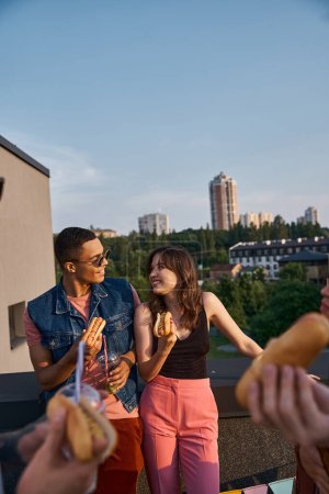 fröhliche multirassische Freunde in lebendigen Outfits genießen köstliche Hot Dogs auf dem Dach bei einer Party