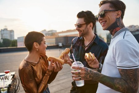 joyous good looking friends with sunglasses in urban outfits enjoying hot dogs at rooftop party