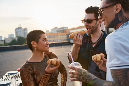 jolly attractive people with stylish sunglasses in vivid outfits enjoying hot dogs at rooftop party