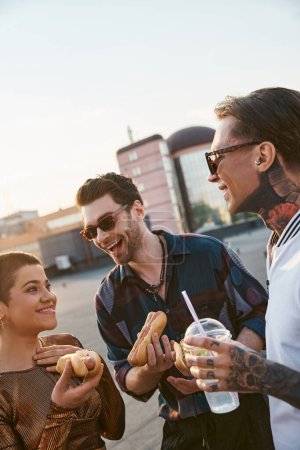 cheerful attractive friends with sunglasses in urban outfits enjoying hot dogs at rooftop party