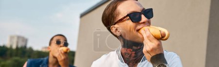 focus on young man with tattoos with his african american friend on backdrop eating hot dogs, banner