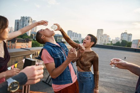 Foto de Jóvenes alegres en trajes elegantes vertiendo tequila en la boca de su amigo afroamericano - Imagen libre de derechos