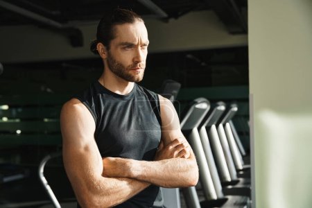 Un hombre atlético en ropa activa se para con confianza frente a una fila de cintas de correr en un entorno de gimnasio.