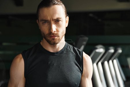 An athletic man in a black shirt stands confidently in front of a row of treadmills in a gym.