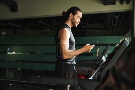 Ein athletischer Mann in aktiver Kleidung steht auf einem Laufband, während er im Fitnessstudio mit seinem Handy trainiert.