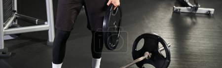 Téléchargez les photos : Un homme se tient avec confiance sur un haltère dans une salle de gym, mettant en valeur l'équilibre et la force. - en image libre de droit