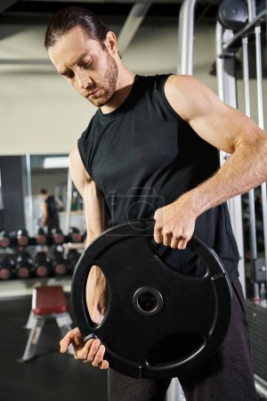 A muscular man in workout attire holds a black plate inside a gym, showcasing strength and determination.